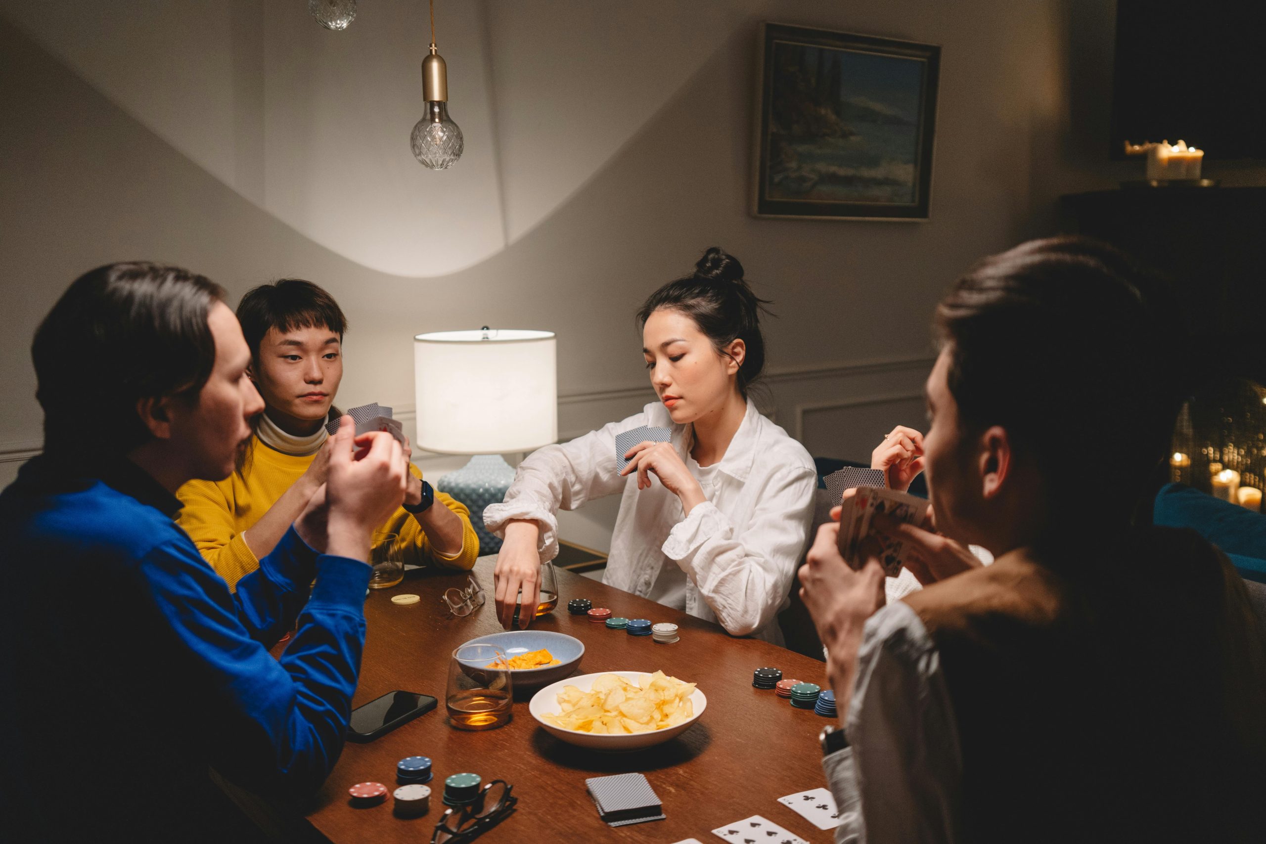 A diverse group of friends playing poker at a wooden table indoors, enjoying snacks and drinks.